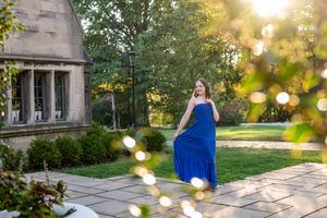 Woman in blue gown walking on stone patio surrounded by lush greenery and golden sunlight.