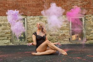 Blonde woman sitting on ground surrounded by colorful pink and purple powder clouds.