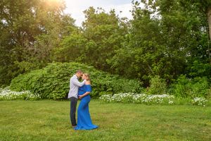 Couple embracing in green park, woman wearing flowing blue maternity gown.