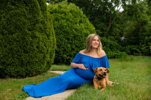 Pregnant woman in a flowing blue off-shoulder gown posing with a brown dog in a lush garden.