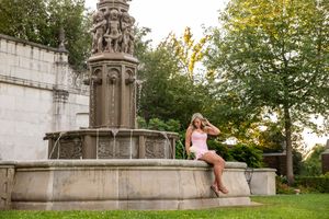 Young woman in pink outfit sitting on edge of ornate stone fountain in lush green park setting.