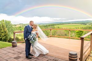 Bride and groom kissing on a wooden deck with a stunning rainbow over lush green vineyard hills in the background.