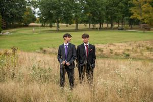 Two young men in dark suits standing in tall golden grass with a lush green golf course backdrop.