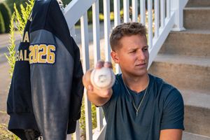 Young man holding a baseball near suburban steps wearing a teal shirt with a varsity jacket hanging nearby.