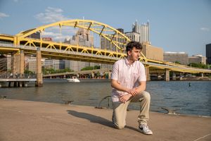 Young man posing by Pittsburgh riverfront with iconic yellow Fort Duquesne Bridge and city skyline in background.