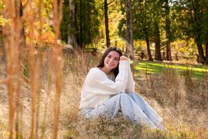 Young woman in white sweater and jeans sitting in autumn field with golden foliage background.