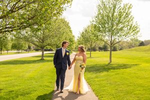 Bride in yellow gown and groom in navy suit walk hand-in-hand along a sunlit park path lined with green trees.