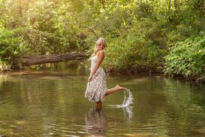 A blonde woman in a floral dress splashes water in a serene forest creek.