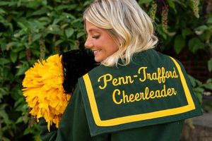 Blonde cheerleader wearing a Penn-Trafford Cheerleader jacket holding a yellow pom-pom outdoors.