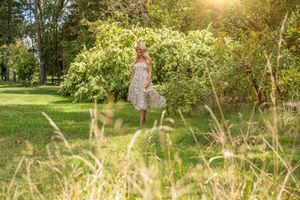Woman in floral sundress standing in lush green park with blooming trees on sunny day.