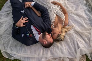 Bride and groom lying on wedding veil outdoors, sharing a romantic kiss on their wedding day.