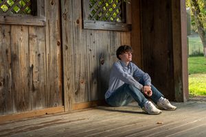 Teenage boy in blue shirt sitting thoughtfully on a rustic wooden porch leaning against barn doors.