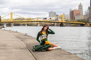 Cheerleader in green uniform posing on riverfront with Pittsburgh city bridge backdrop.