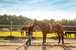 Young woman standing with a saddled brown horse at a sunny ranch with horses grazing in the background.