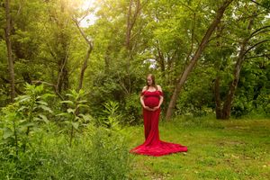 A pregnant woman in a red gown poses gracefully in a lush green forest.