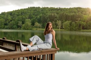 Young woman in white tank and gray pants sitting on a wooden dock overlooking a serene lake.