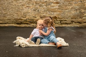 Two young toddlers sitting on a blanket outdoors, sharing a sweet moment with a stone wall background.