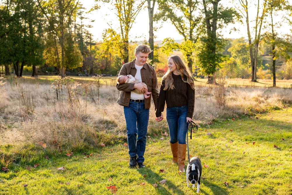 Young couple walking with newborn baby and dog through a sunny autumn meadow.