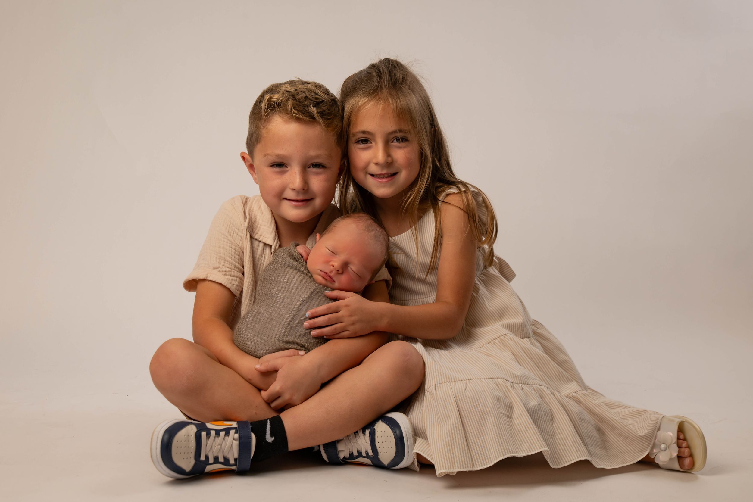 Two children smiling while gently holding a sleeping newborn baby in a white studio.