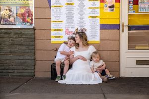 A bride in a white dress sits with two young children outside a shop.