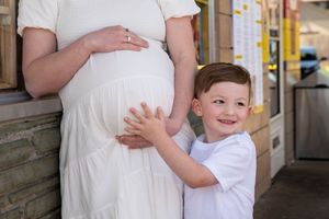 Smiling young boy touching pregnant mothers belly outdoors on sunny day.