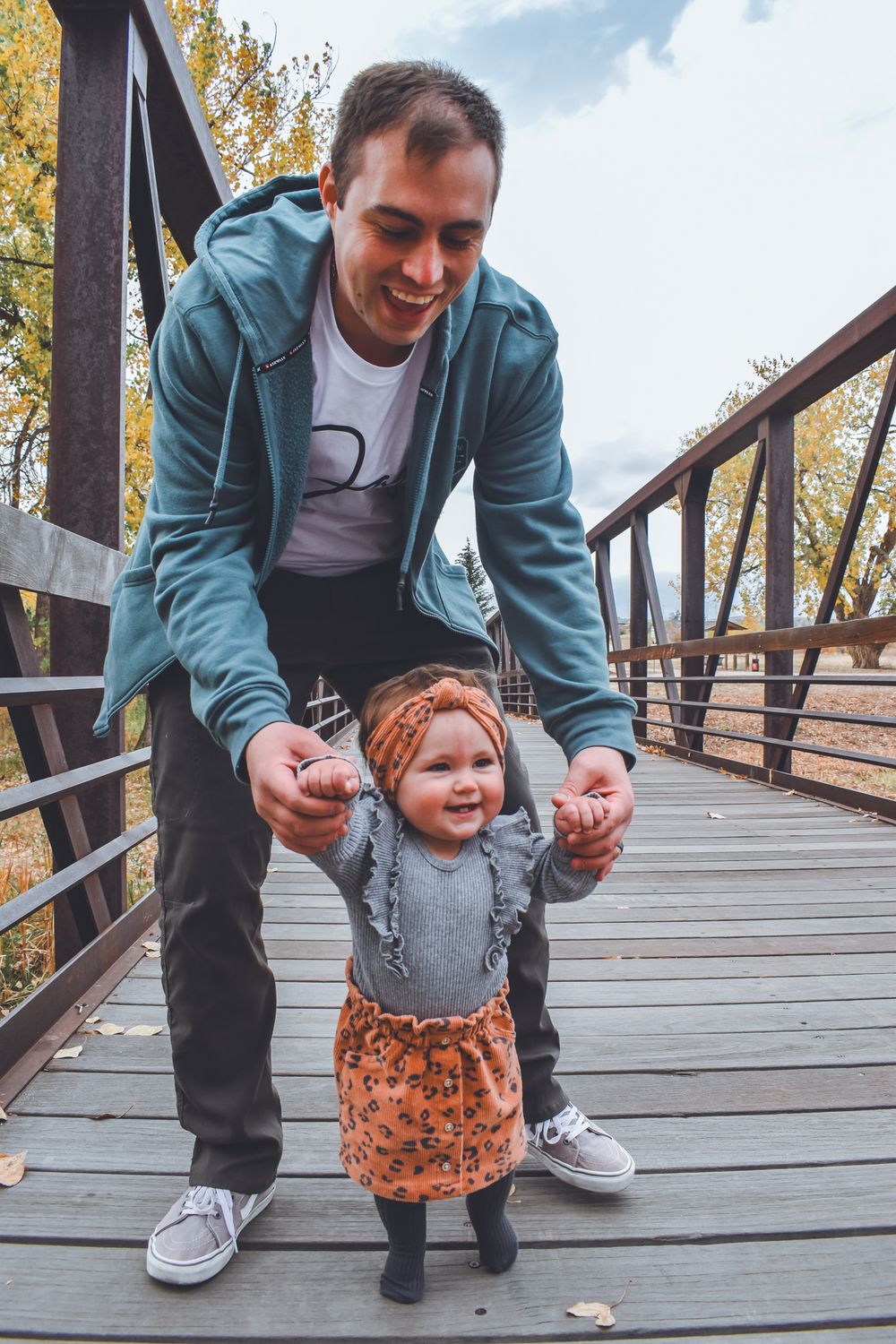 Smiling father helping his baby daughter stand on a wooden bridge surrounded by fall foliage.