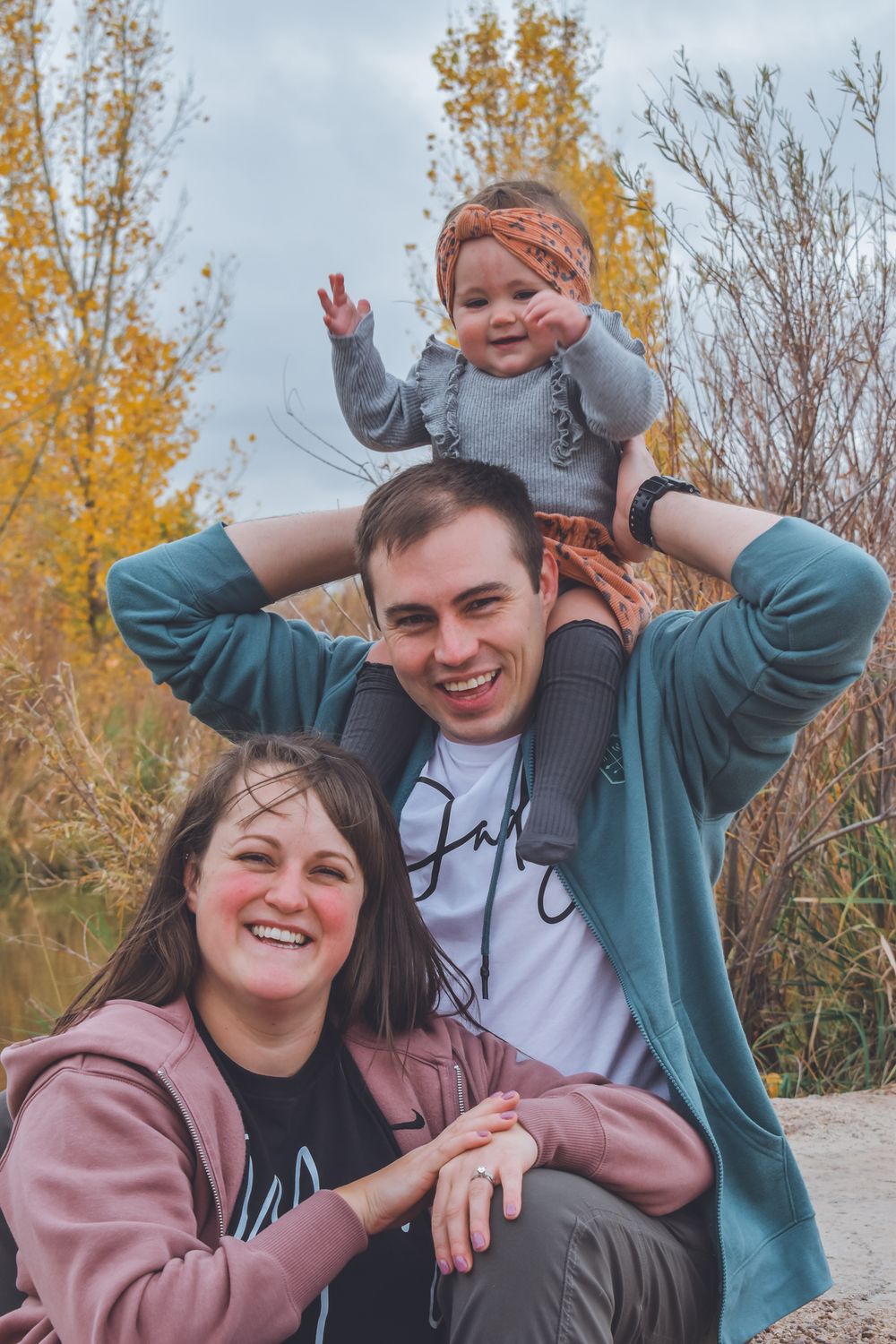 A happy young family with a baby on dad's shoulders poses outdoors among autumn foliage.