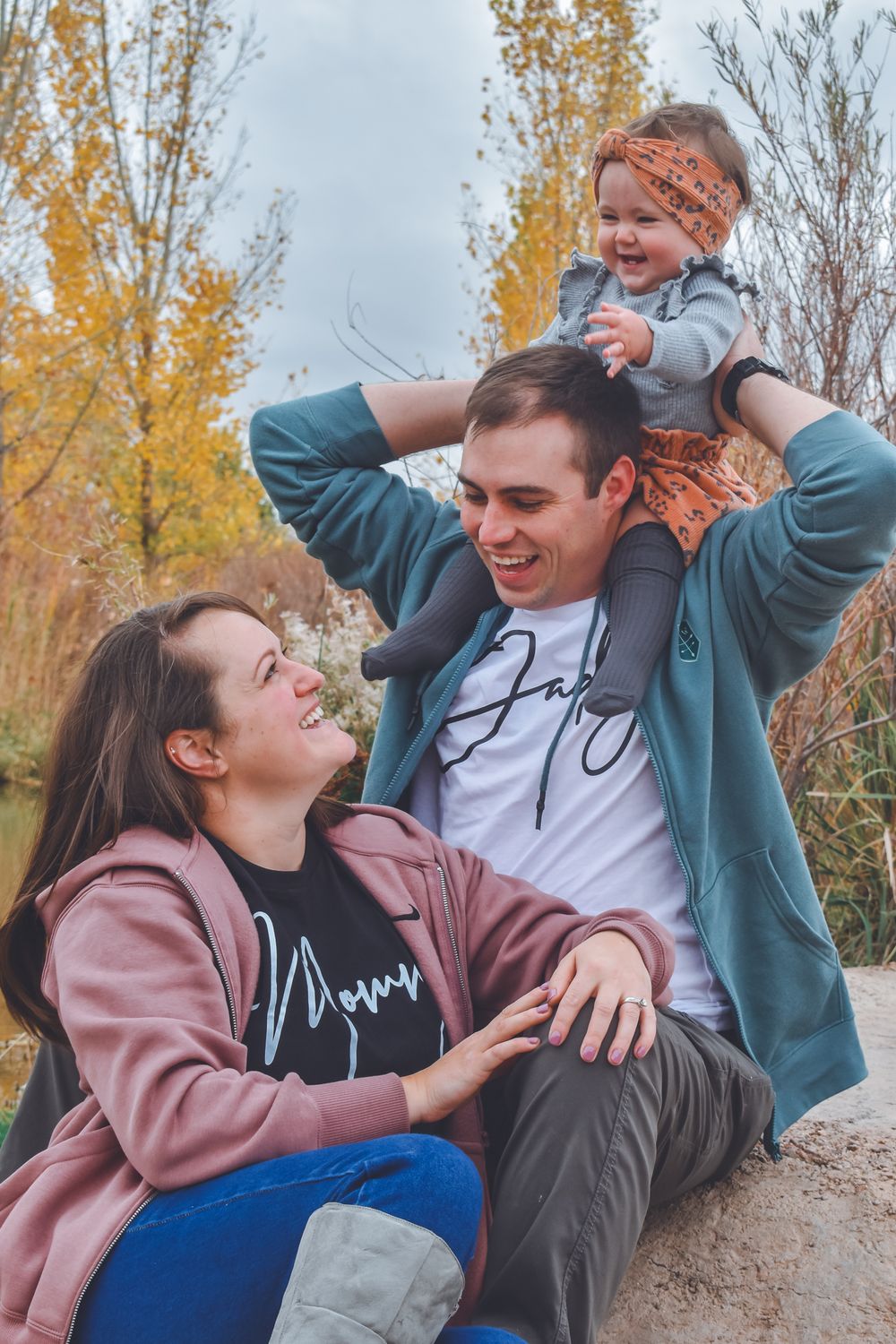 Joyful family of three with father lifting baby on shoulders while mother laughs in autumn setting.
