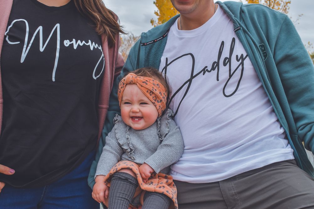 A couple wearing matching Mommy and Daddy shirts sit together holding their smiling baby girl in an outdoor fall setting.