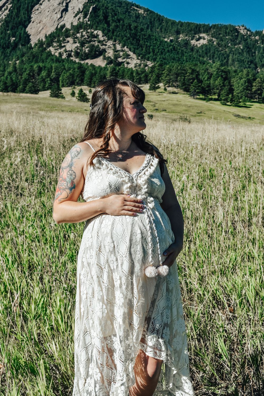 Pregnant woman in white lace dress holding baby bump in a scenic mountain meadow.