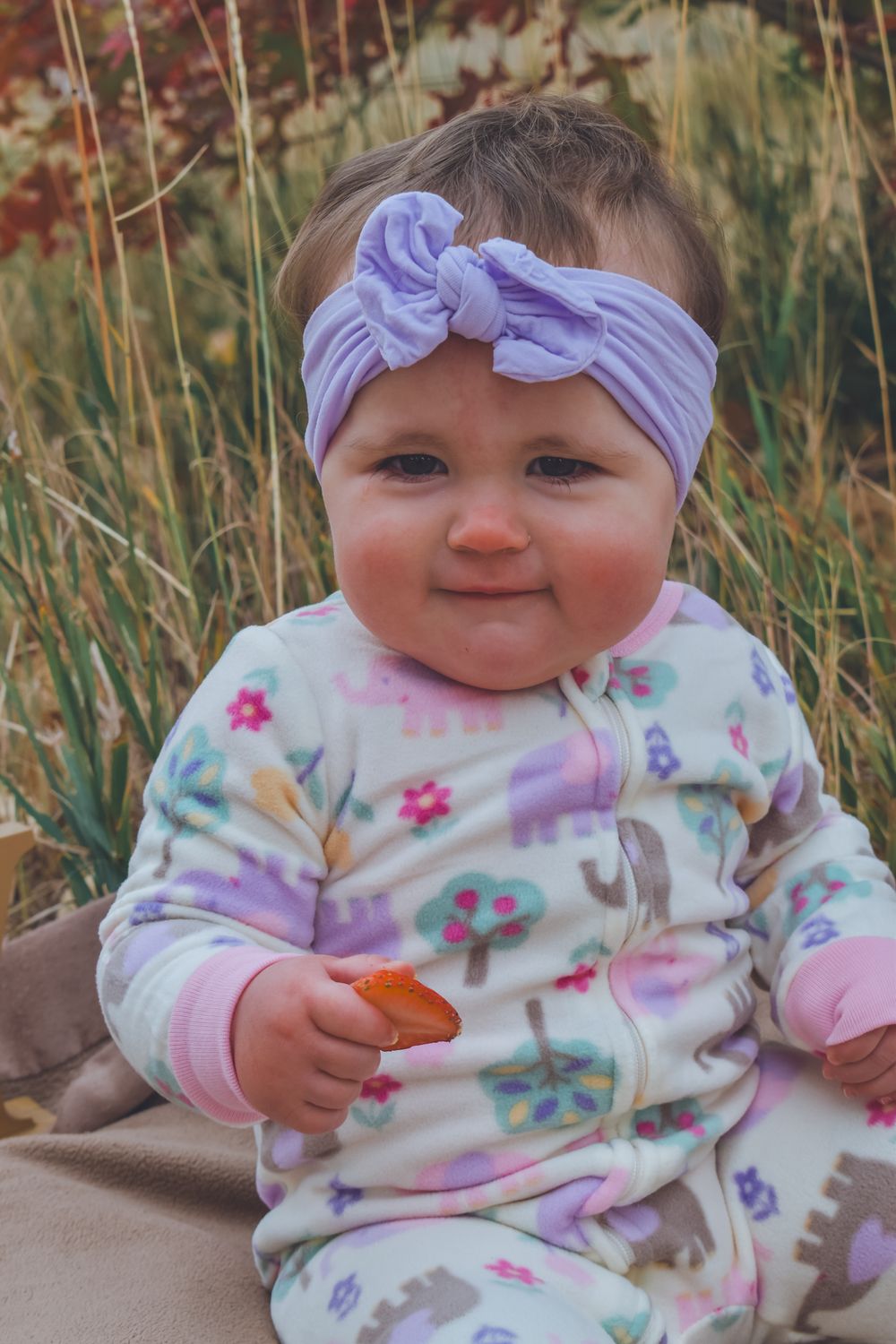Adorable baby girl wearing purple bow headband and floral onesie sitting outdoors holding a small snack.
