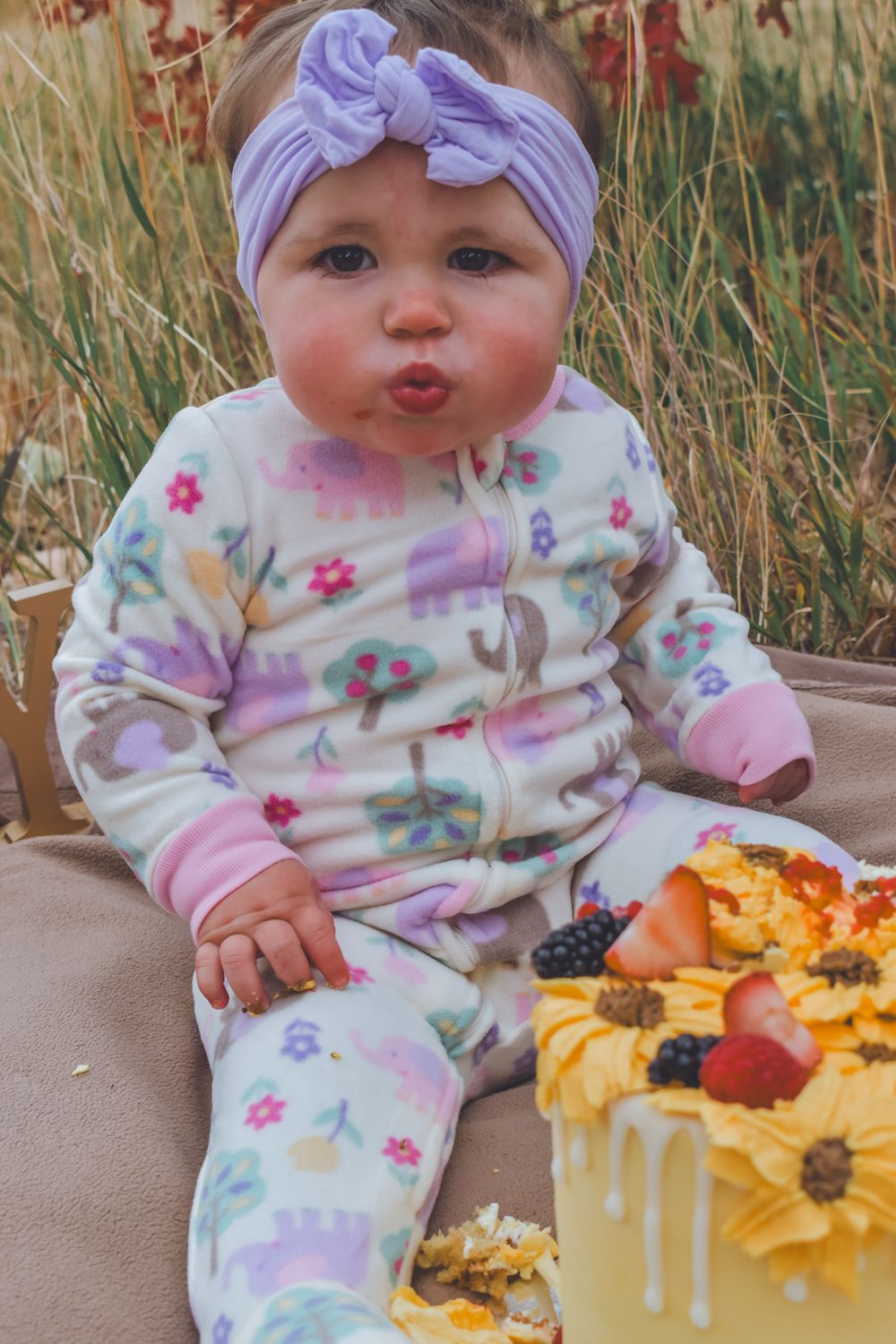 Cute baby girl in floral pajamas with purple bow headband sitting beside yellow birthday cake.
