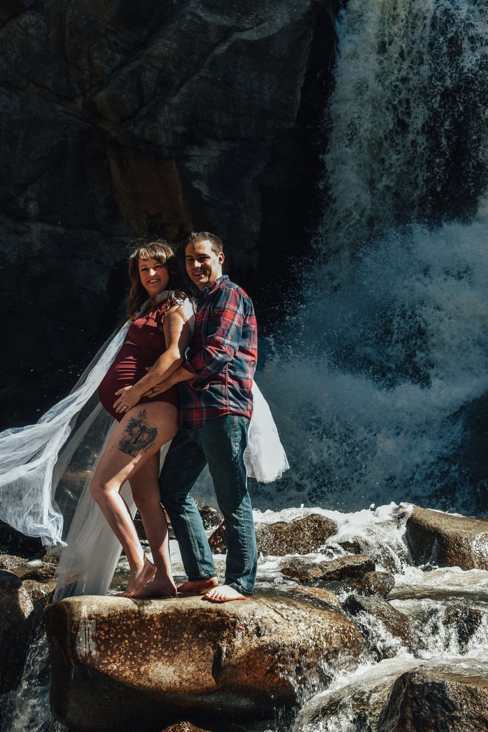 Couple embracing on rocks in front of a waterfall, woman in red dress with veil.