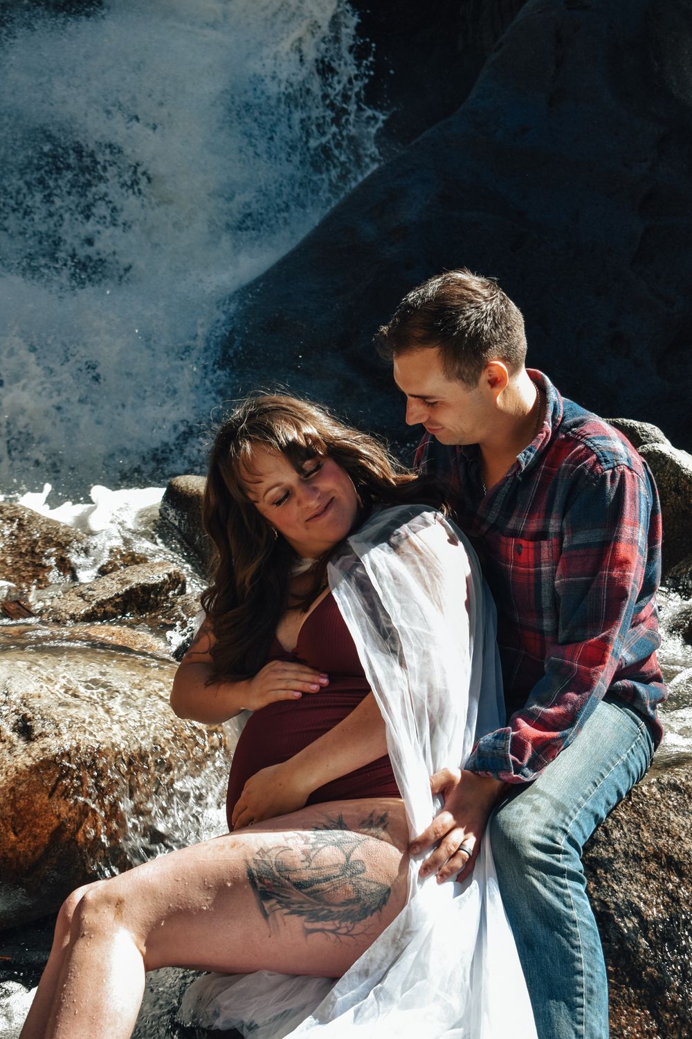 Couple sharing a tender moment by a waterfall, pregnant woman in burgundy dress with flowy white fabric outdoors.