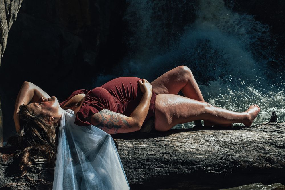 Pregnant woman in red dress lying on rocks near a waterfall, showcasing maternity photography outdoors.