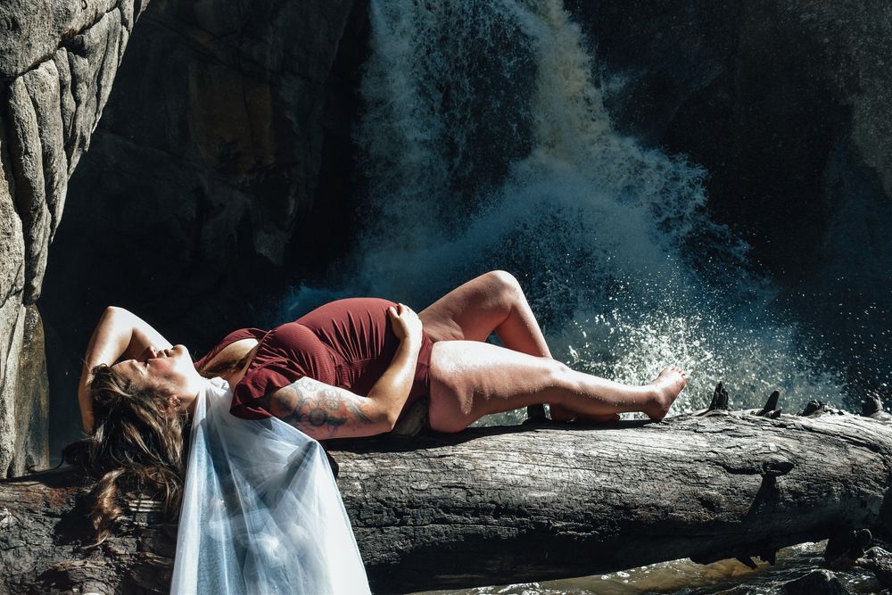 Pregnant woman in burgundy dress lying on a log with a waterfall in the background outdoors.