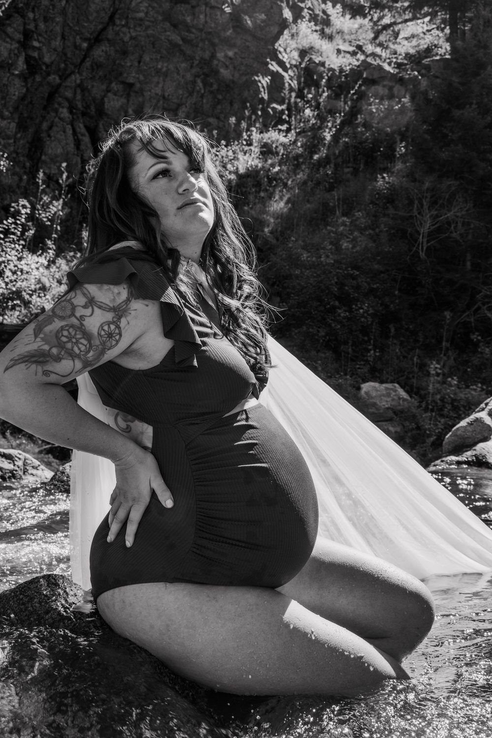 Black and white photo of a pregnant woman with tattoos sitting on rocks near a stream in a nature setting.