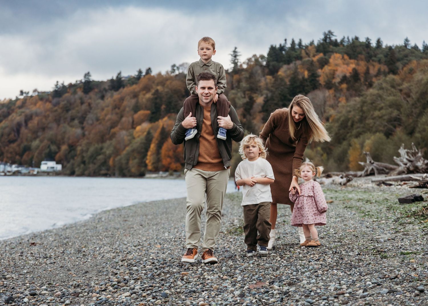 A family enjoys a stroll together along a rocky beach shore with forested hills in the background.