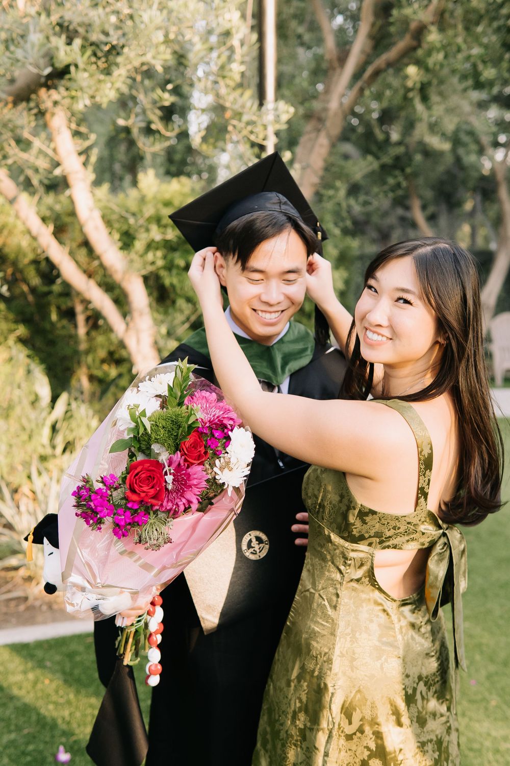 Smiling Asian couple at graduation, woman in gold dress adjusting mans black cap while holding colorful flower bouquet outdoors.