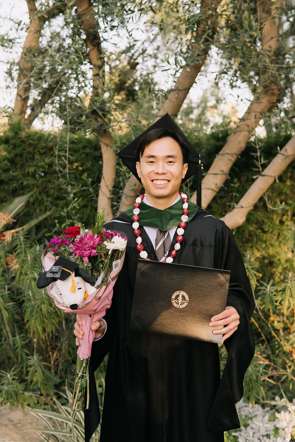 Smiling graduate in black cap and gown holding flowers and diploma outdoors.