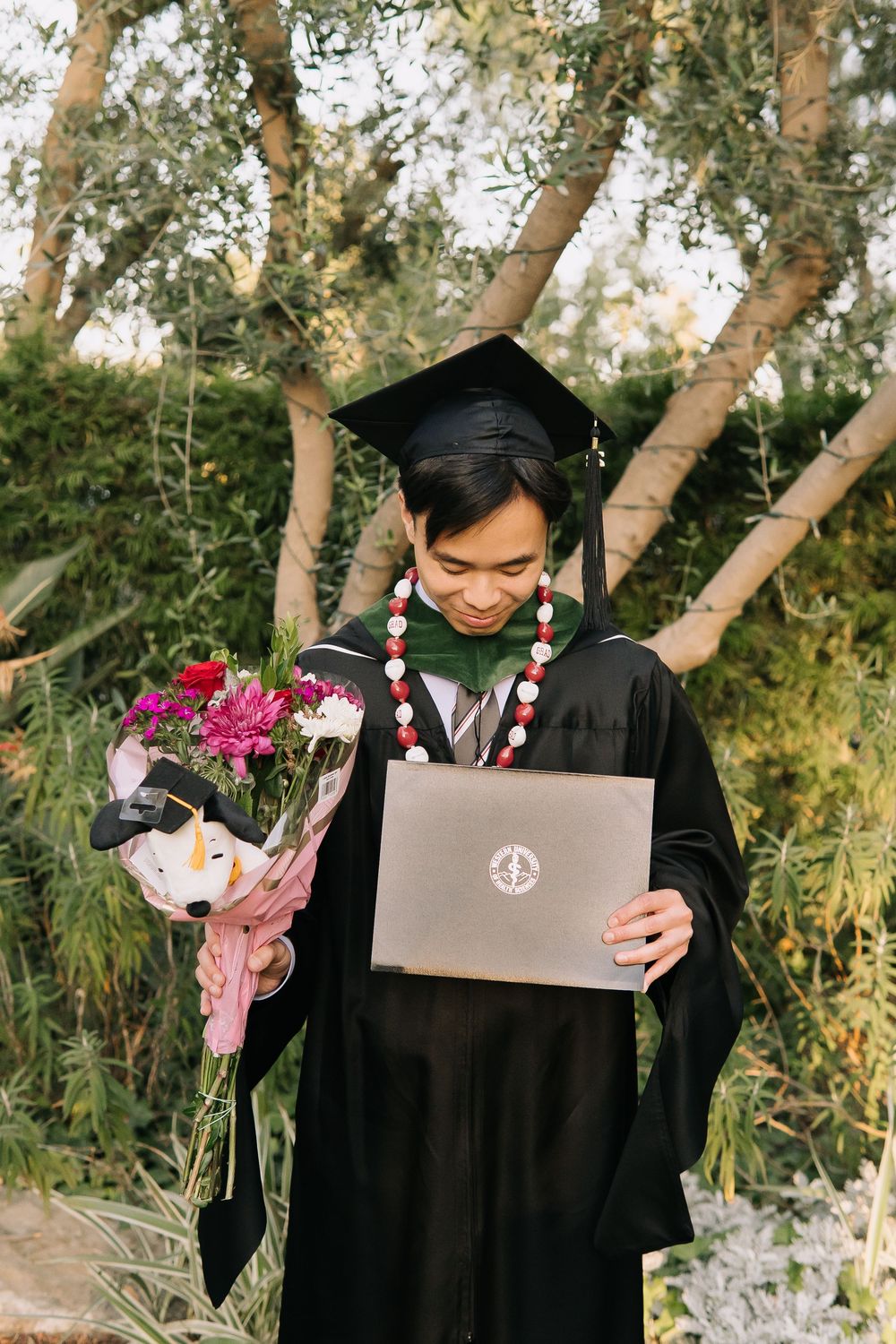 Young man in graduation cap and gown holding diploma and flowers, smiling outdoors among green trees.
