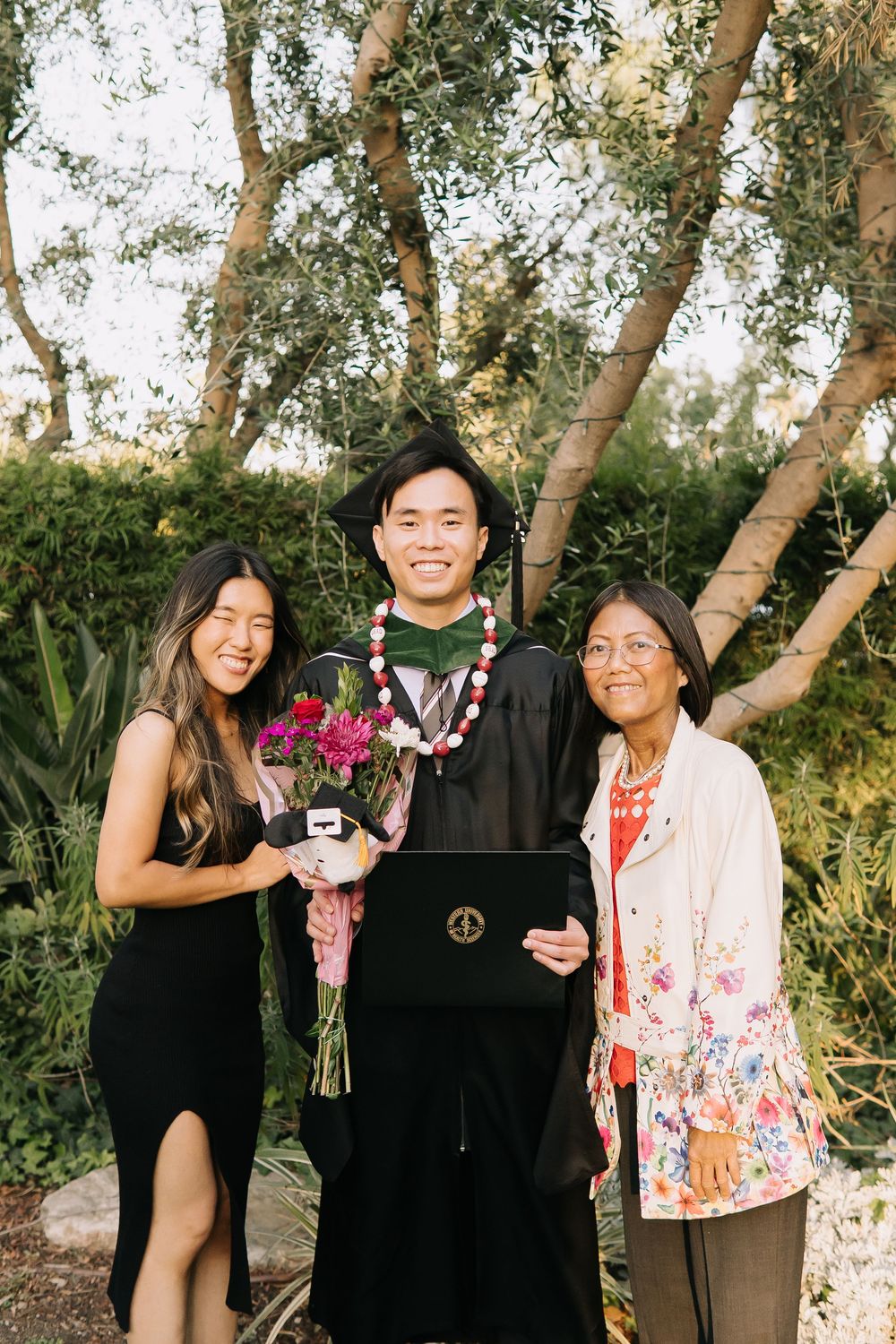 Graduate in cap and gown holds diploma and flowers, posing with two smiling women outdoors near lush green trees.