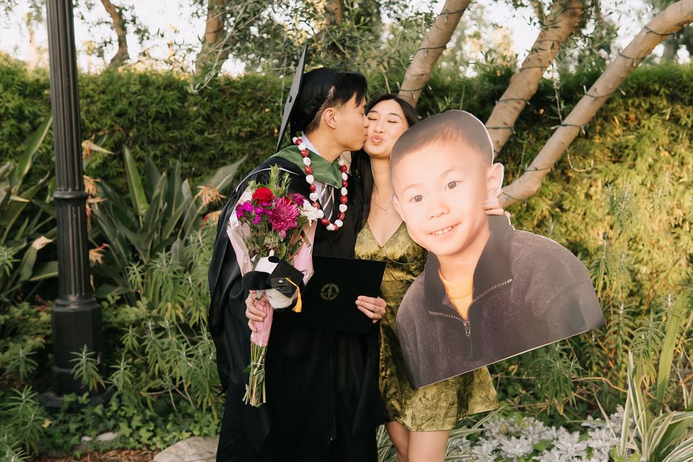 Graduate in cap and gown kisses partner while holding flowers, with a childhood photo cutout in a garden setting.