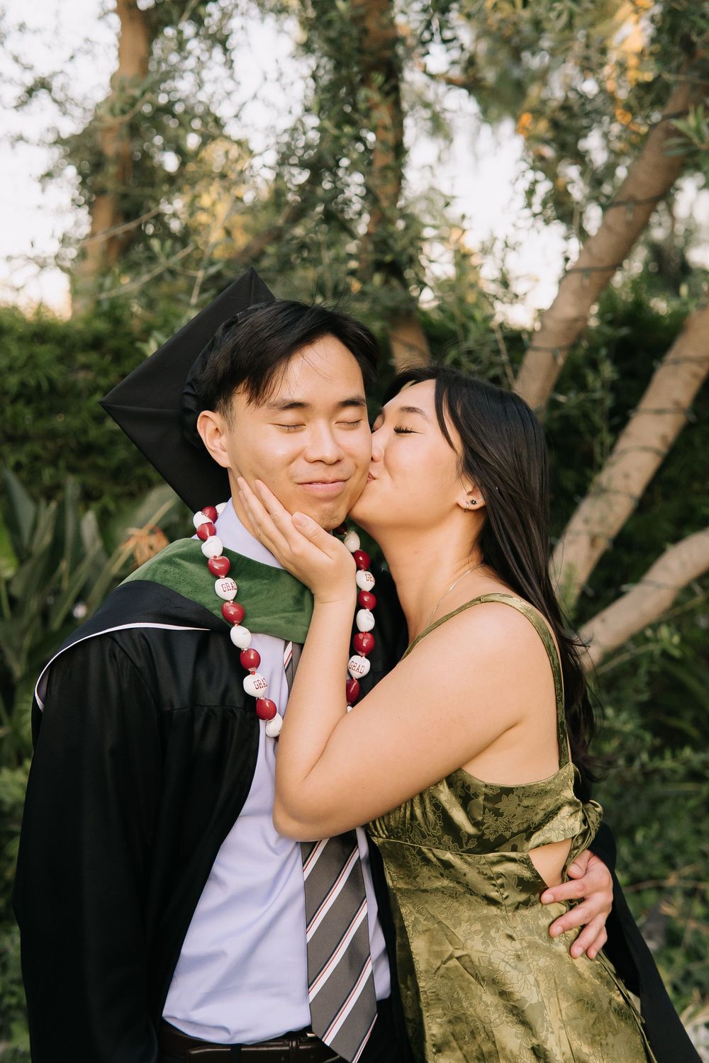 A woman in a green dress kisses a man in graduation cap and gown wearing a lei outdoors.