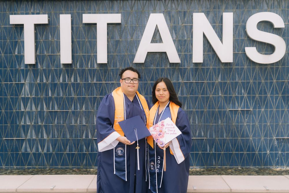 Two graduates in blue and gold robes pose proudly in front of a Titans wall at their graduation ceremony.