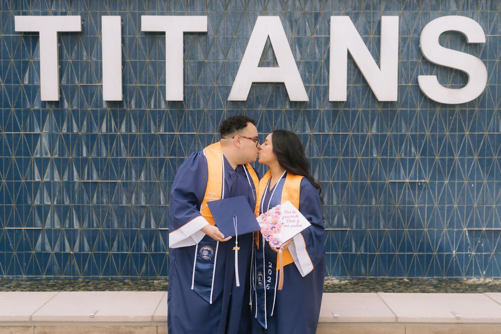 Two graduates in blue robes share a kiss in front of a Titans wall during their graduation celebration.