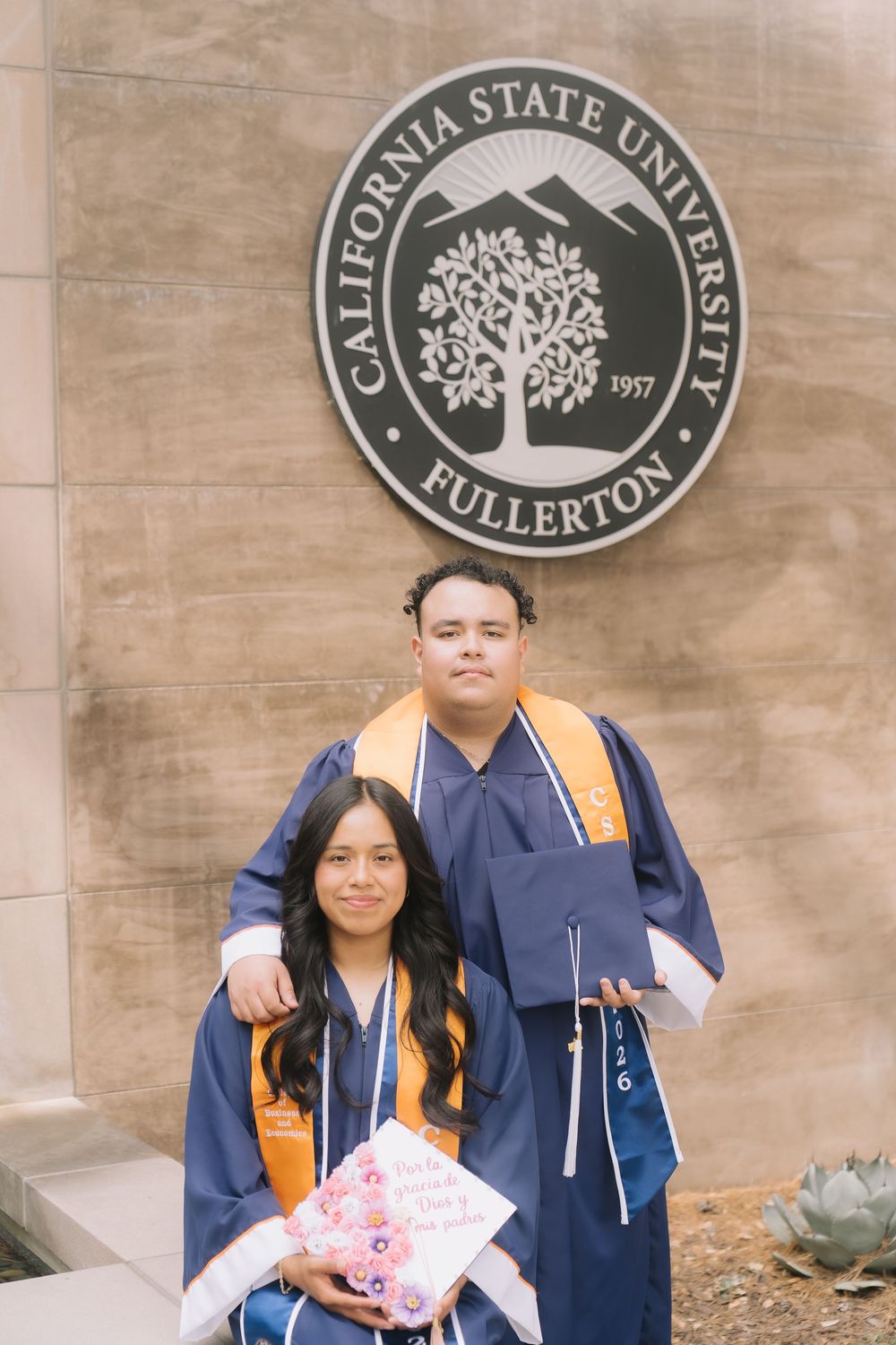 Two graduates in blue robes and gold stoles pose proudly in front of California State University Fullerton seal.