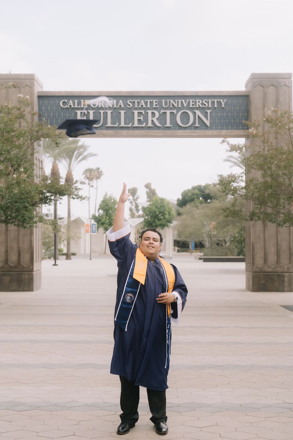 Graduate in blue cap and gown tossing hat at California State University Fullerton entrance arch.