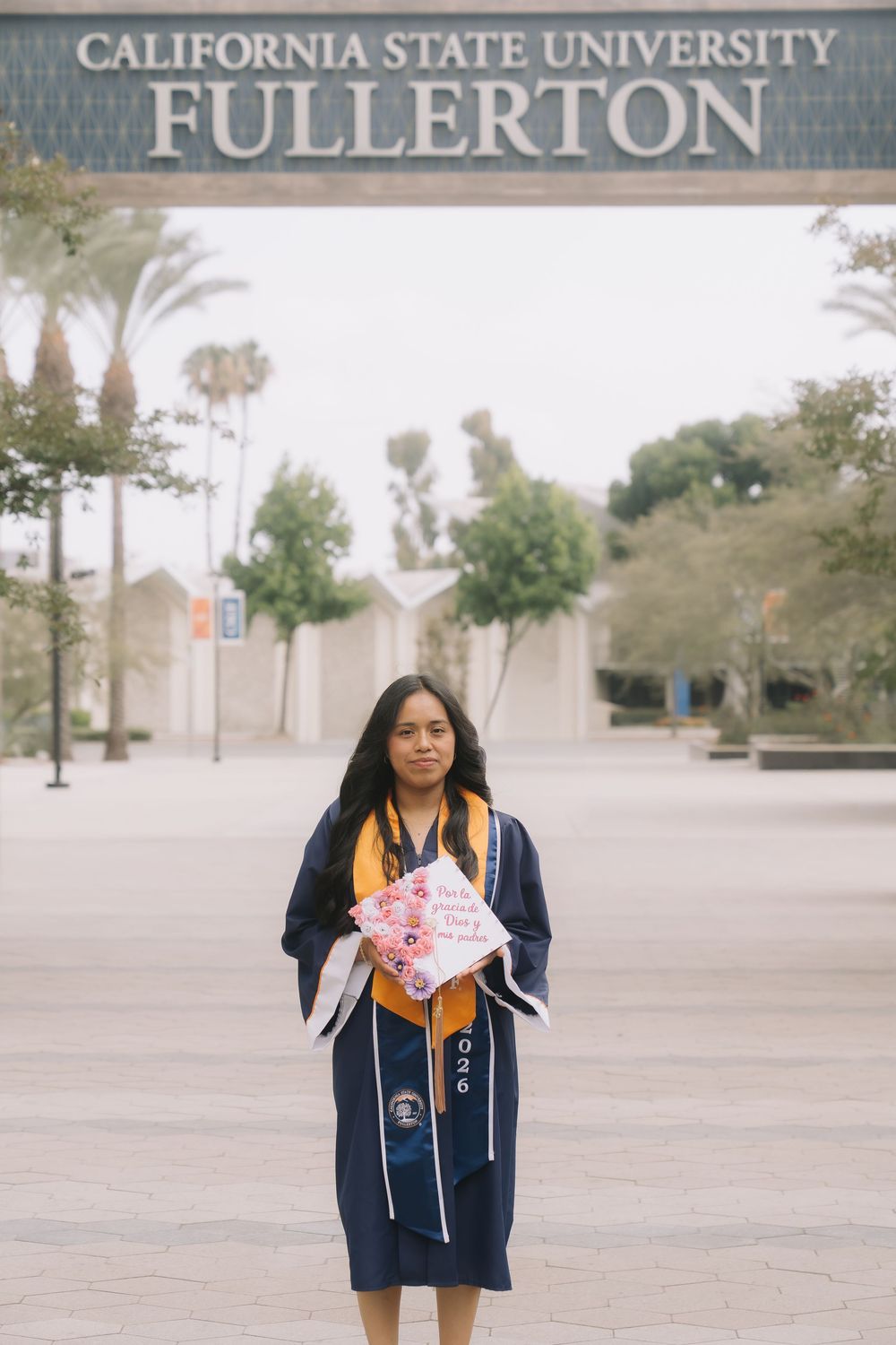 A graduate in cap and gown holds a decorated mortarboard in front of California State University Fullerton sign.