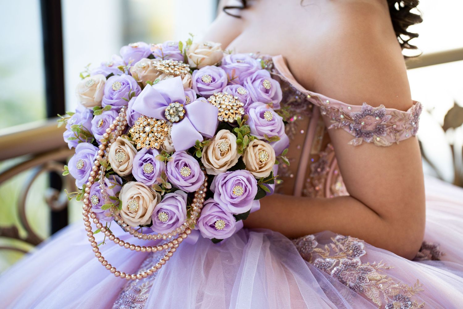 A bride holds a lavender and cream rose bouquet with gold pearl accents, wearing an off-shoulder lilac ballgown.